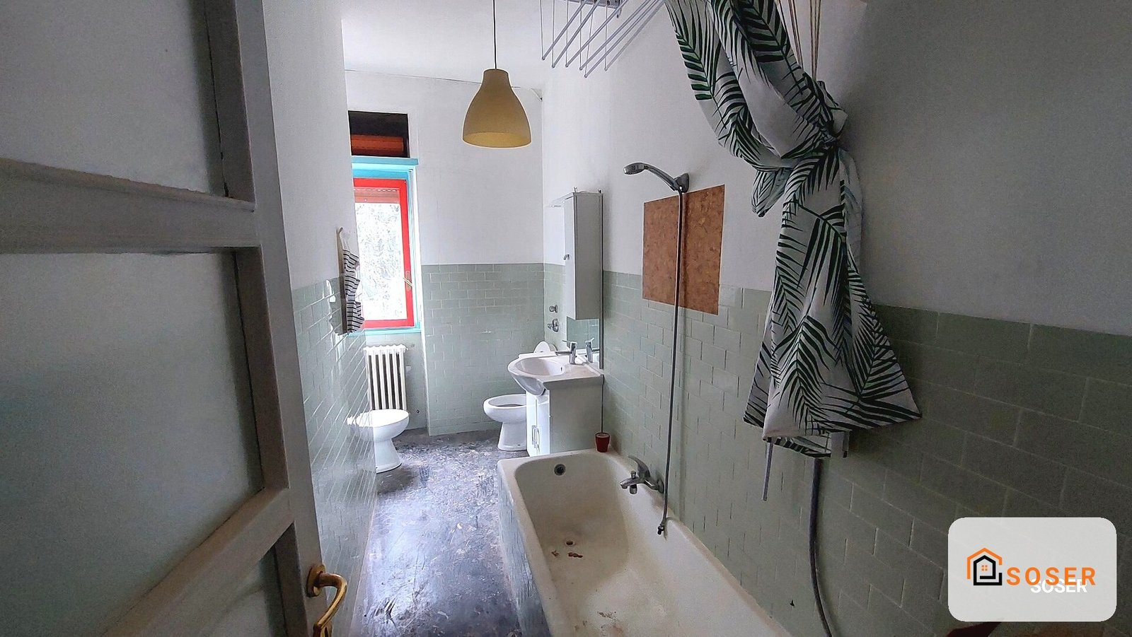 Bagno in ristrutturazione con vecchie piastrelle verdi, vasca, lavabo e finestra con cornice rossa