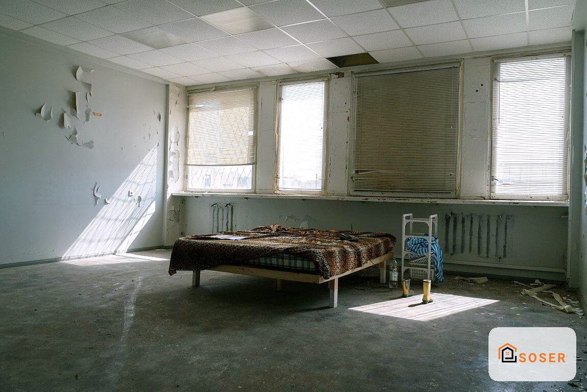 Camera da letto abbandonata con letto singolo, pareti scrostate e finestre con veneziane, da ristrutturare