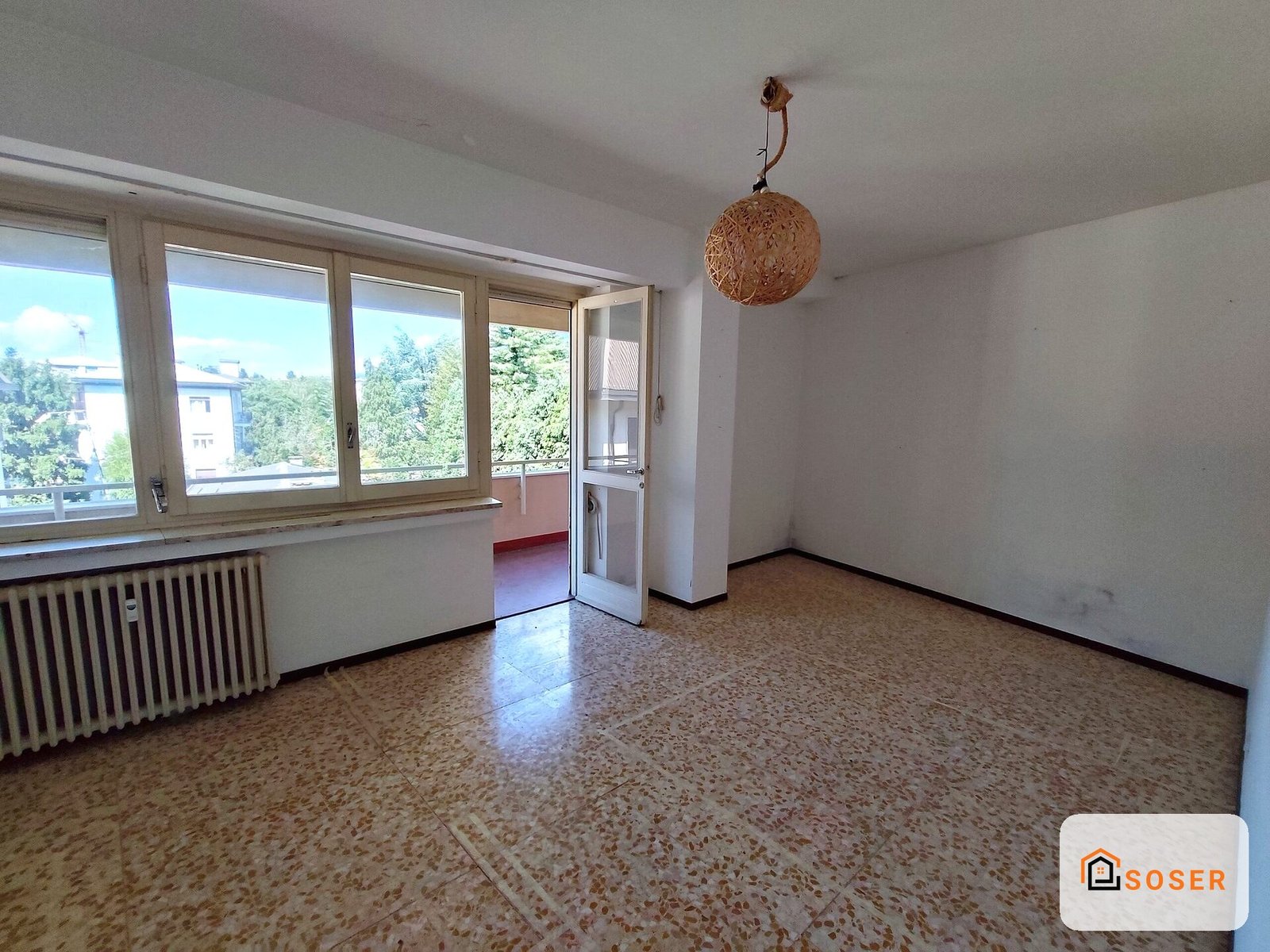 Soggiorno vuoto con pavimento in terrazzo, finestre in legno e balcone, prima della ristrutturazione