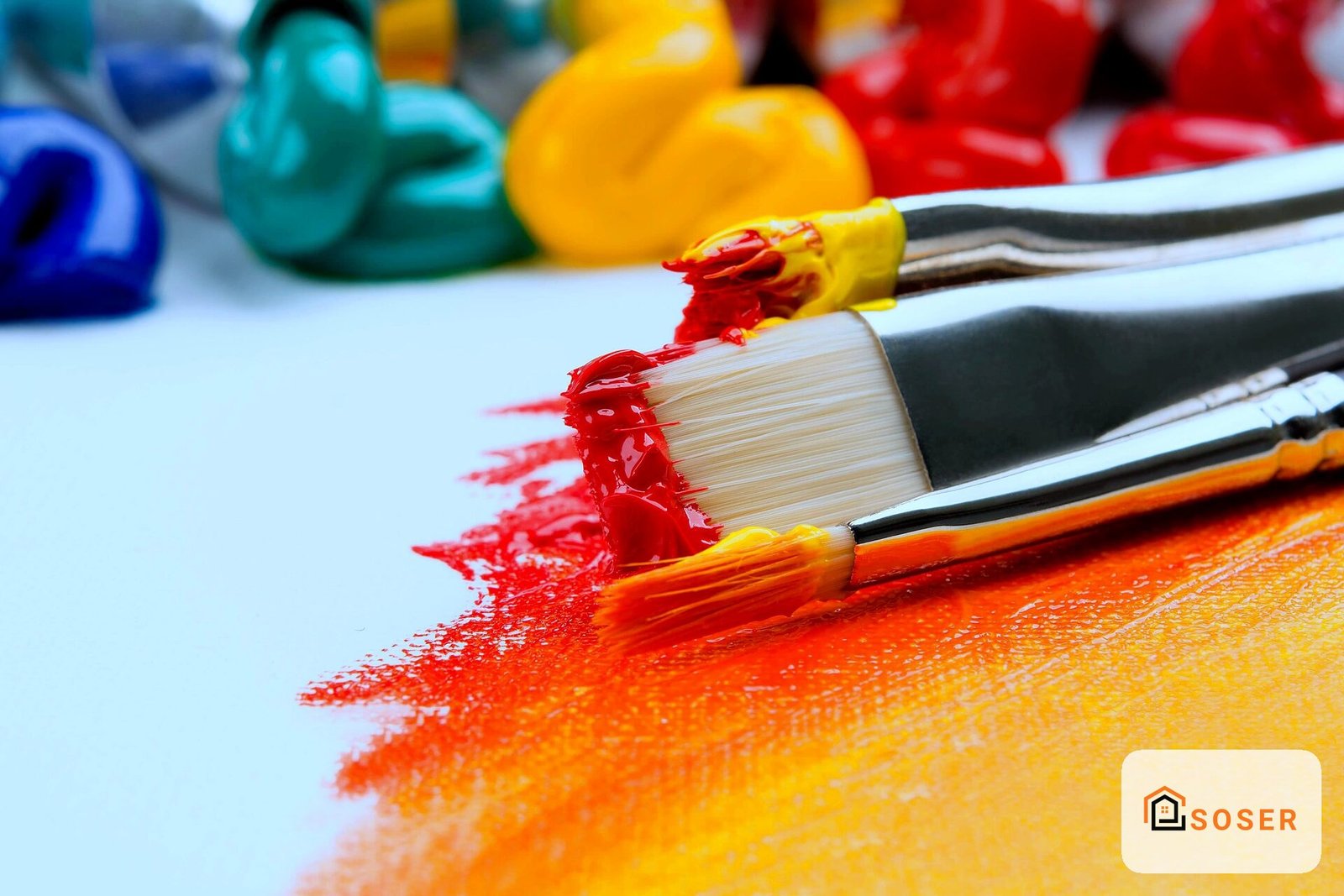 Pennelli con vernice rossa e gialla su superficie bianca con palette di colori sfocata sullo sfondo