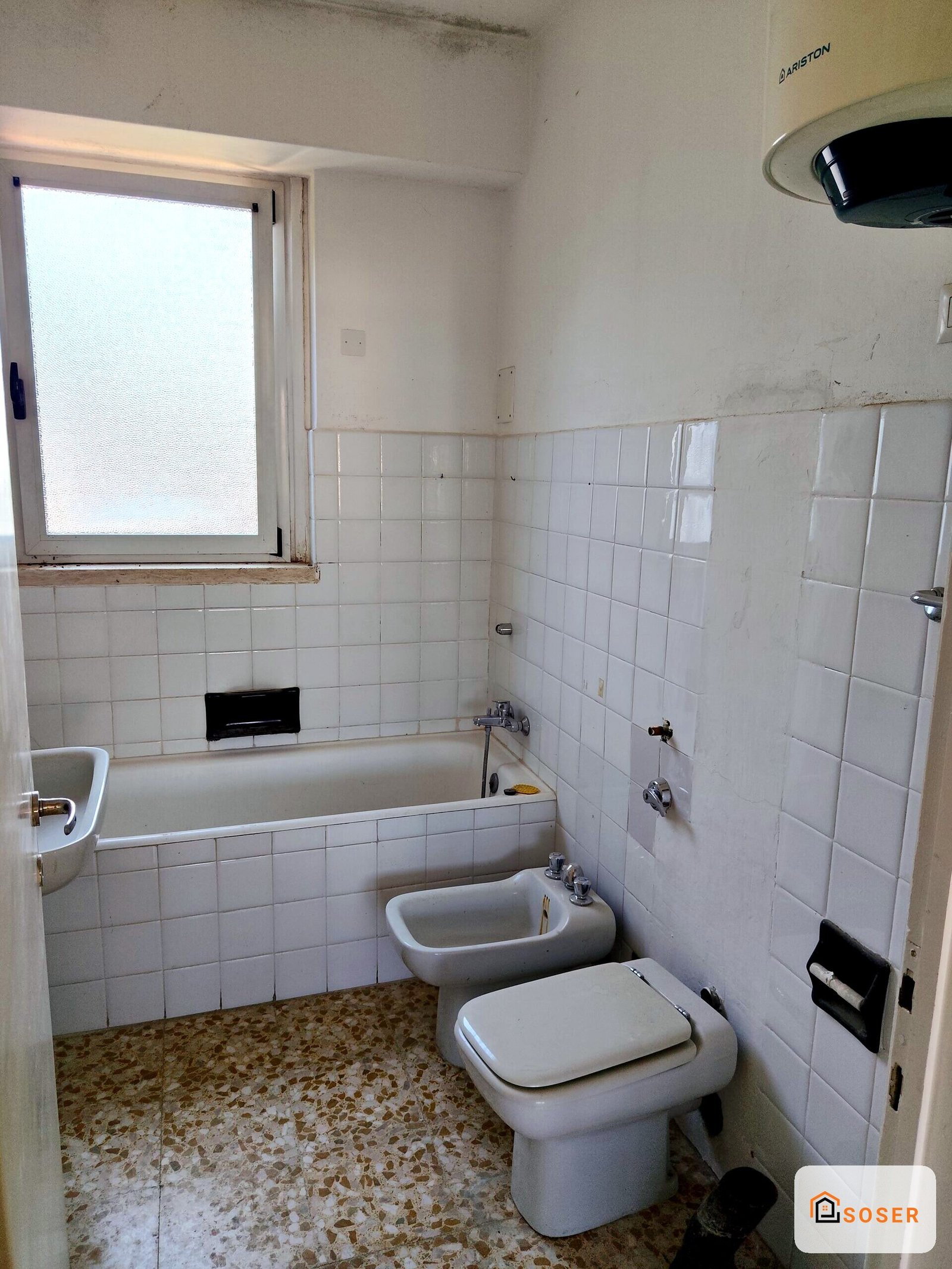Bagno datato con piastrelle bianche e pavimento in terrazzo, toilette e bidet bianchi, vasca con rivestimento