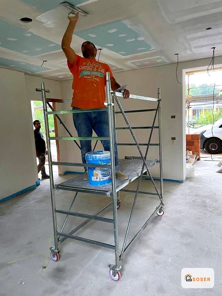 Operaio su scaffale mobile dipinge il soffitto in cartongesso di interno in ristrutturazione