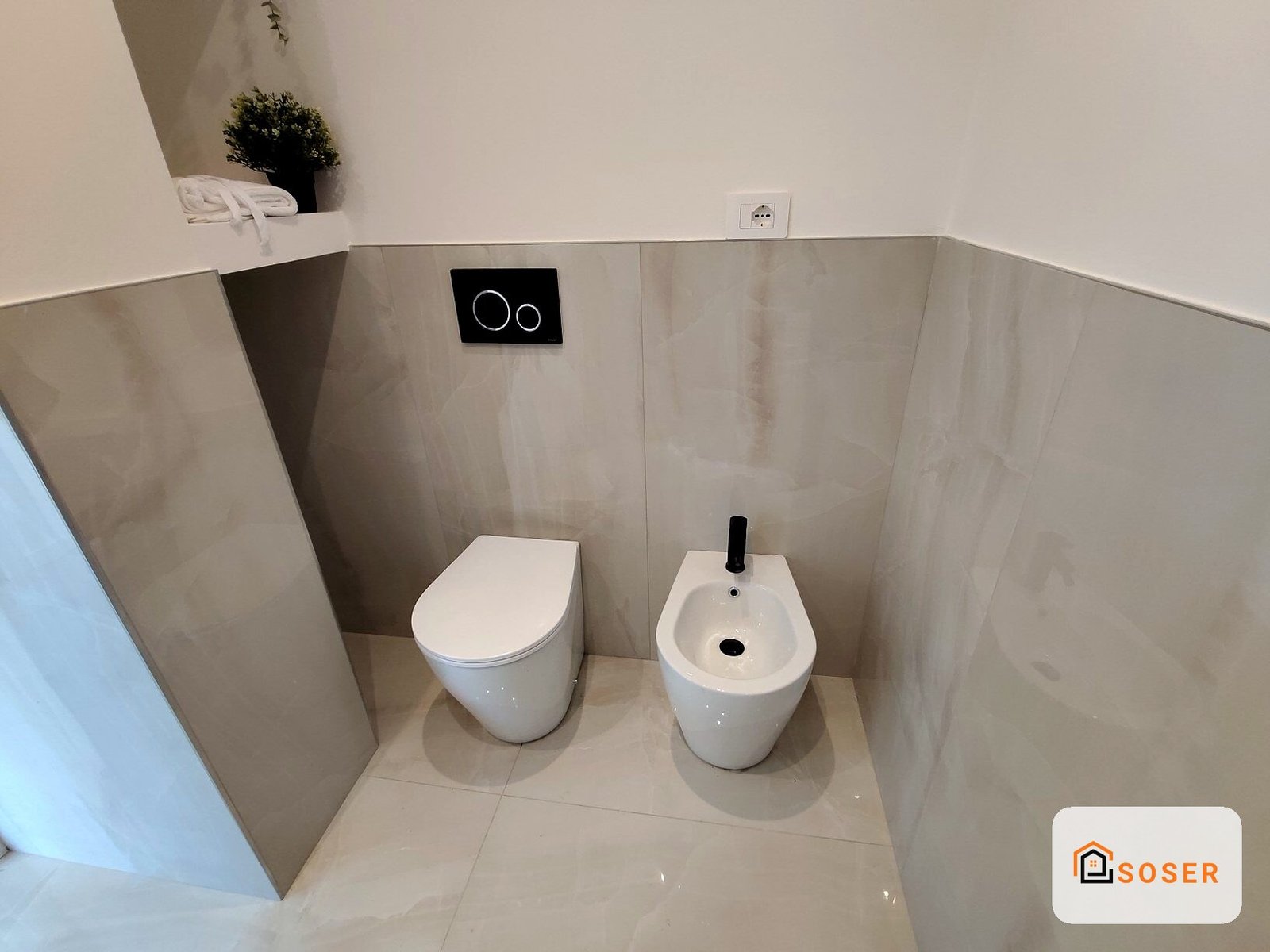 Bagno moderno con wc e bidet bianchi, rivestimento in piastrelle beige e nicchia con pianta