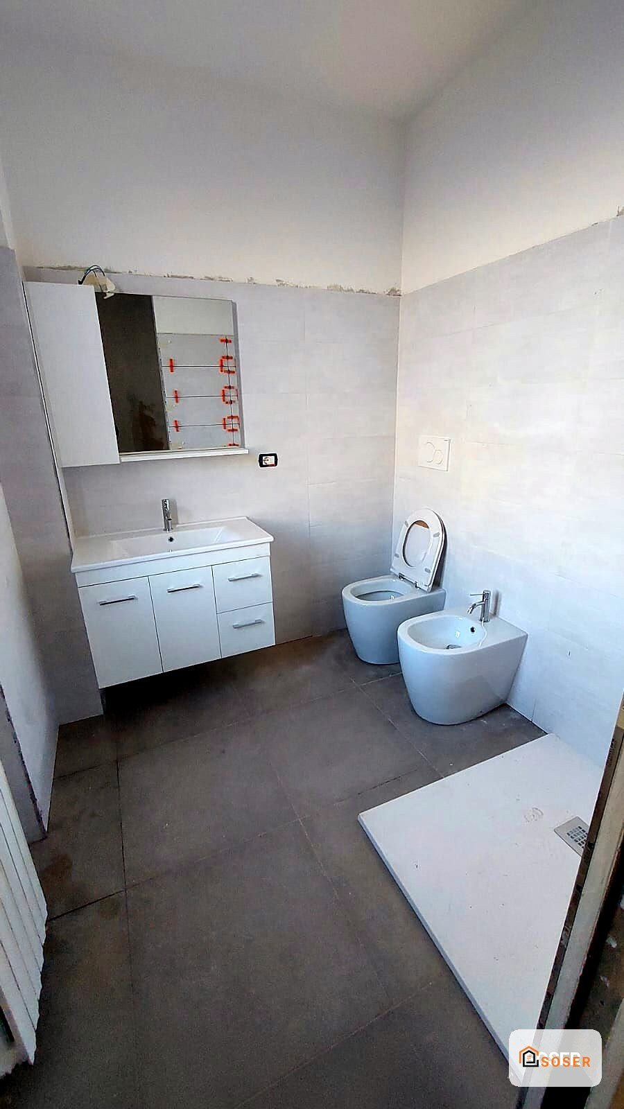 Bagno in ristrutturazione con sanitari bianchi moderni, lavabo sospeso e specchio, pavimento grigio