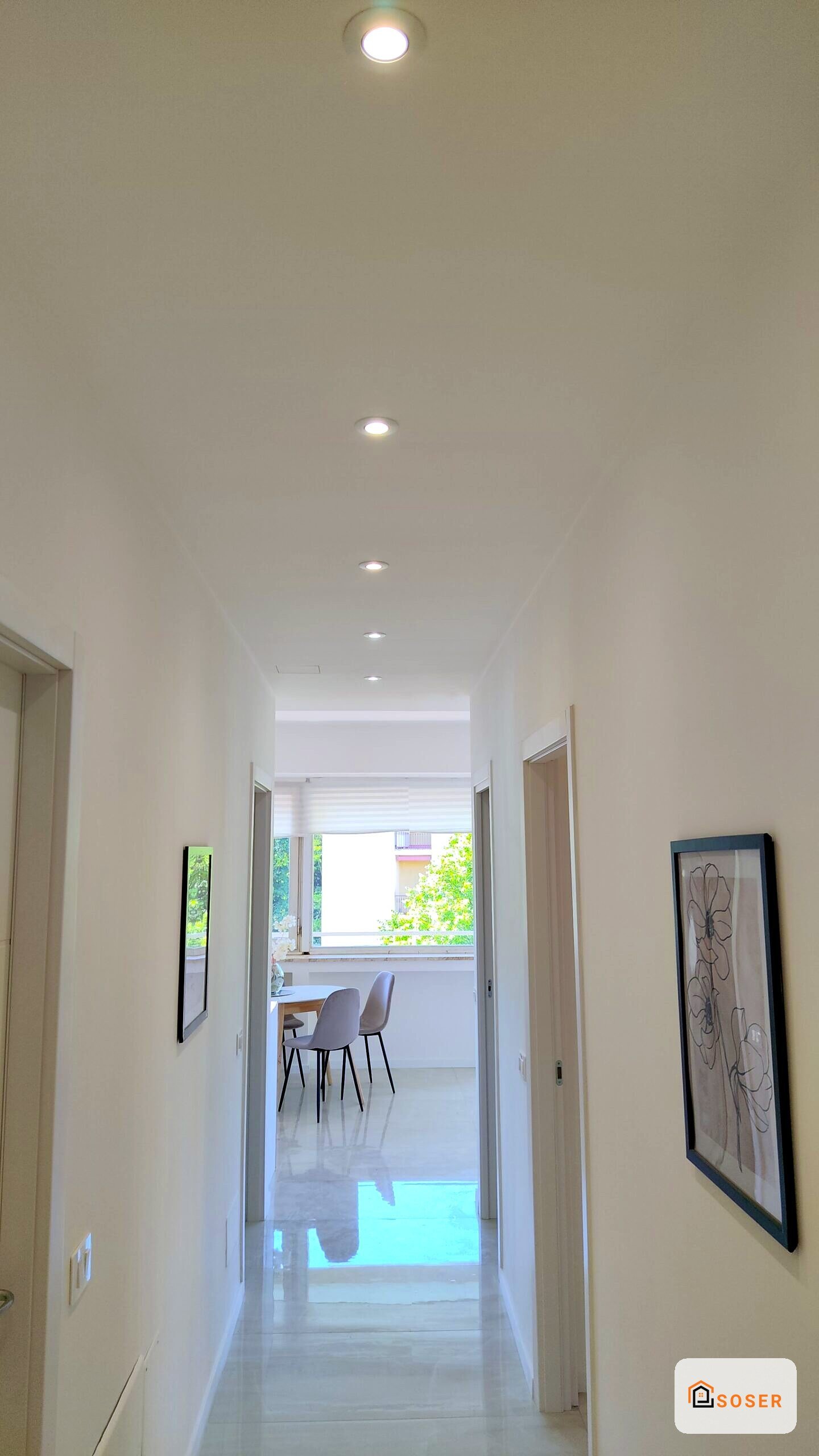 Corridoio moderno con illuminazione a incasso, pareti bianche e pavimento lucido che conduce a una zona living luminosa