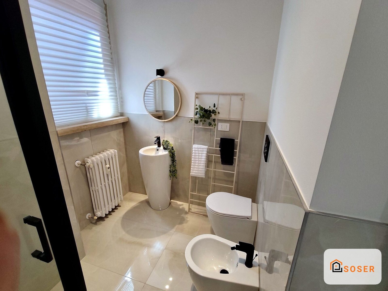Bagno moderno ristrutturato con lavabo bianco, specchio rotondo, radiatore e piastrelle beige